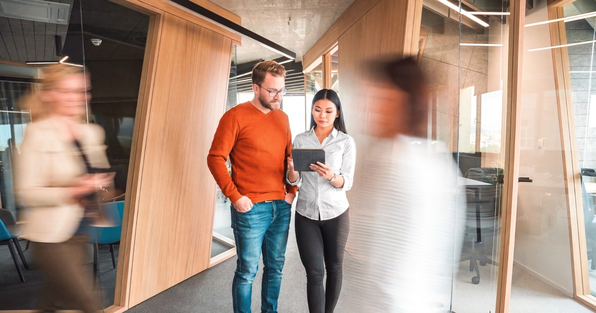 Man and woman in office look at tablet while walking and other walk by