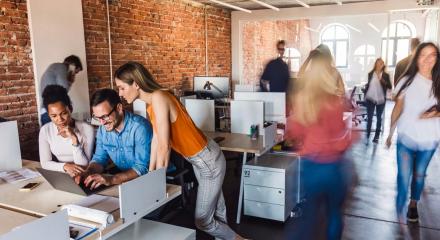 Three people in office look at screen around desk while others walk by